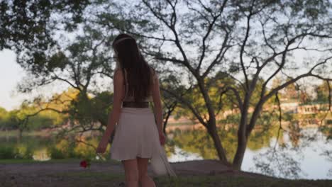 Beautiful-woman-walking-carefree-with-hat-and-rose-in-her-hands-near-lake-with-vegetation-at-sunset