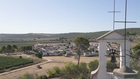 Aerial-footage-captures-La-Ermita-del-Poblado-de-San-Julián,-adorned-with-metal-crosses-and-a-bell-tower,-against-the-backdrop-of-the-quaint-residential-area-of-San-Julián