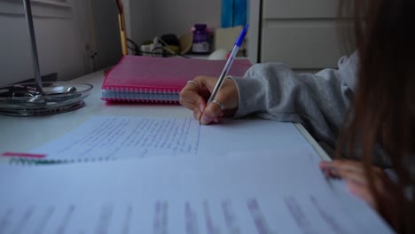 Unrecognizable-young-girl-student-taking-notes-with-blue-pen-on-desk-by-window