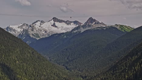 Whistler,-BC,-Kanada,-Luftaufnahme-Eines-Bewaldeten-Berges-Mit-Einer-V8-Drohne,-Die-Die-Fahrt-Einer-Gondelbahn-Durch-Grüne-Täler-Mit-Schneebedeckten-Gipfeln-Im-Hintergrund-Einfängt-–-Aufgenommen-Mit-Mavic-3-Pro-Cine-–-Juli-2023