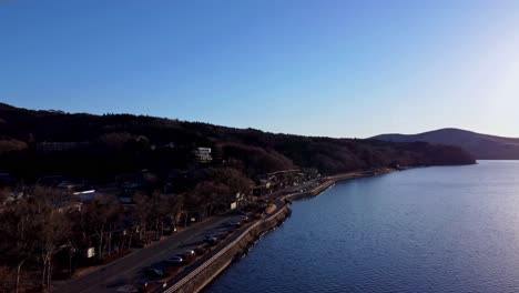Coastal-road-curving-along-a-calm-bay-with-hills,-clear-sky-at-dusk,-aerial-view