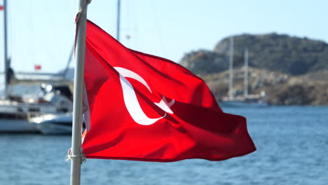 Turkish-Flag-Waving-With-The-Wind-With-Boat-And-Sea-In-The-Background-In-Gumusluk,-Turkey