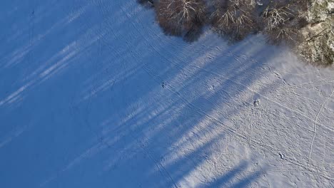 Ein-Weites,-Schneebedecktes-Feld,-Das-In-Sonnenlicht-Getaucht-Ist,-Mit-Langen-Schatten,-Die-Sich-Von-Unsichtbaren-Objekten-über-Die-Landschaft-Erstrecken,-Aufgenommen-Aus-Der-Luftperspektive-An-Einem-Klaren-Wintertag