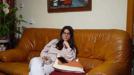 Mujer-Con-Gafas-Relajándose-En-Un-Sofá-Marrón-Viendo-La-Televisión,-Tomando-Un-Sorbo-De-Un-Vaso