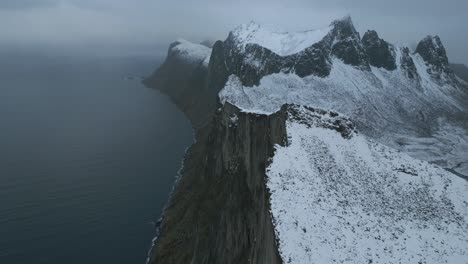 Snow-covered-Hesten-mountain-on-Senja-Island,-Norway,-with-dramatic-cliffs-overlooking-the-sea,-under-overcast-skies