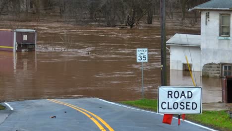 Road-closed-sign-in-front-of-flooded-river-with-brown-murky-water-on-street