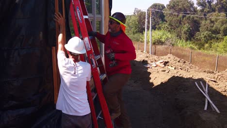 Gimbal-booming-up-shot-of-a-construction-worker-ascending-a-ladder-and-securing-the-siding-of-a-pre-fabricated-house-at-a-modular-construction-site-in-Los-Angeles,-California