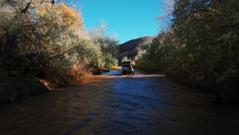 Rear-view-of-white-SUV-driving-through-shallow-creek-bed-with-fall-foliage,-Utah