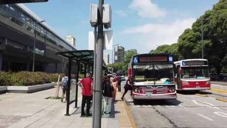 Der-Rote-Autobus-Kommt-Im-Sommer-Bei-Tageslicht-Am-Bahnhof-Belgrano-In-Buenos-Aires-An,-Einem-Wahrzeichen-Des-öffentlichen-Nahverkehrs-In-Argentinien,-Einheimische-Und-Fußgänger,-Die-Warten