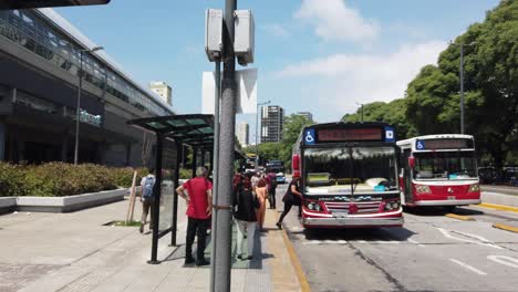 El-Autobús-Rojo-Llega-A-La-Estación-De-Belgrano-En-La-Ciudad-De-Buenos-Aires,-Argentina,-Un-Punto-De-Referencia-Del-Transporte-Público-A-La-Luz-Del-Día-De-Verano,-La-Gente-Local,-Los-Peatones-Esperando