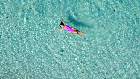 A-woman-in-a-pink-swim-dress-swims-in-the-transparent-Mediterranean-Sea,-viewed-from-above