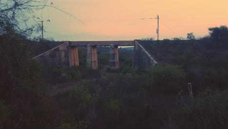 Toma-Aérea-De-Un-Antiguo-Puente-Ferroviario-De-Hormigón-Abandonado-Con-Densas-Colinas-Forestales-En-El-Fondo-Durante-La-Tarde