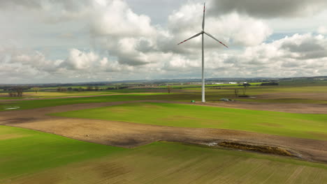 Rising-drone-shot-of-idyllic-green-farm-fields-and-rotating-Wind-turbine-against-cloudy-sky