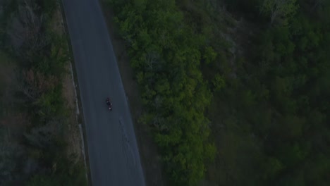 Aerial-shot-in-plan-of-motorbike-traveling-along-an-empty-road-in-the-hills-with-camera-tilt-up-on-mountains