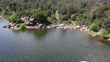 Flug-Im-Rückzug-Und-Aufstieg-In-Einem-Reservoir,-Visualisierung-Des-Ufers-Mit-Felsen-Und-Klarem-Wasser-Und-Entdeckung-Eines-Waldgebiets-Mit-Kiefern-Und-Tannen-An-Einem-Frühlingsmorgen-In-Avila,-Spanien