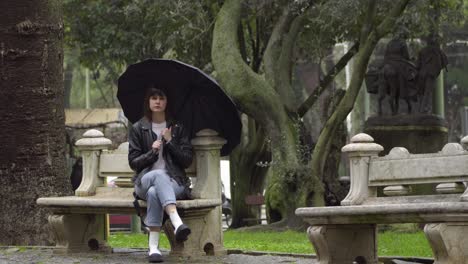 Sad,-frustrated-young-woman-sitting-on-the-bench-in-the-park-under-black-umbrella