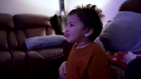 An-18-month-old-baby-boy-watches-TV-in-the-evening,-captivated-by-the-screen-and-clapping-his-hands-in-approval-with-a-curious-expression,-showcasing-the-concept-of-mimicking-behavior-in-slow-motion