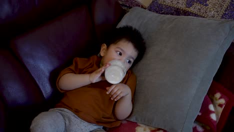 Adorable-18-month-old-baby-boy-sits-leisurely-on-a-home-sofa,-holding-a-bottle-of-milk-and-drinking-from-it,-bathed-in-the-warm-glow-of-TV-screen-light-reflections