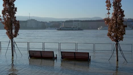 Bancos-Y-árboles-Inundados-En-El-Muelle-De-Budapest-Con-Un-Barco-Navegando-Al-Fondo-Durante-La-Inundación-Del-Río-Danubio,-Hungría---28-De-Diciembre-De-2023