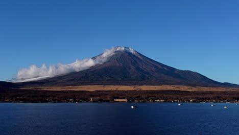 Majestic-Mount-Fuji-with-snowy-peak,-clear-blue-sky-and-lake-in-the-foreground,-serene-nature-scene