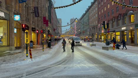 Blustery-flurries-fly-as-pedestrians-cross-snowy-street-Kungsgatan-with-holiday-decorations-in-Stockholm,-Sweden