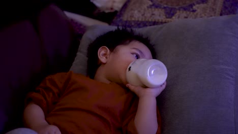 Niño-Asiático-Británico-Con-Camisa-Naranja-Acostado-Bebiendo-De-Una-Botella,-Centrado-En-La-Pantalla-Del-Televisor,-En-El-Interior