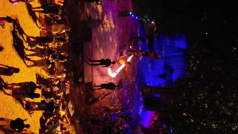 Blonde-girl-jumping-fire-rope-at-Thai-beach-bar-amid-tourist-crowd,-VERTICAL