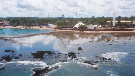 Aerial-view-of-Praia-do-Forte-beach,-the-coral-reef,-palm-tree-area-and-the-clouds-reflecting-on-the-ocean,-Praia-do-Forte,-Bahia,-Brazil