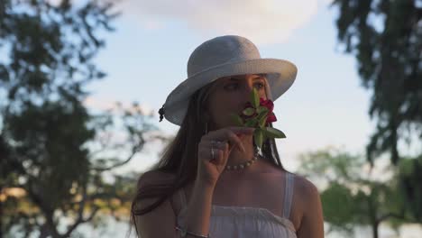 Beautiful-woman-with-hat-carefree-smelling-rose-surrounded-by-vegetation-at-sunset