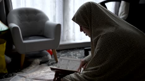Seated-on-the-floor-and-covered-with-a-pile-of-clothing,-a-young-muslim-girl-reads-a-religious-book,-reflecting-the-idea-of-studying-and-learning-within-the-comfort-of-home