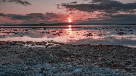 Dramatic-sunset-sky-with-red-sun-and-dark-clouds-above-the-calm-fjord-at-low-tide-in-a-timelapse-video