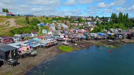 Drone-Aéreo-Sobre-Pilotes-Pueblo-Colorido-En-La-Isla-Mar-Chiloé-Patagonia-Chilena-Destino-De-Viaje-Estableciendo-Tiro,-Colinas-De-Islotes-Verdes-Con-Océano-Pacífico-Azul