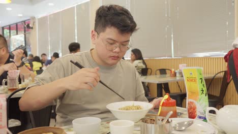 Hombre-Asiático-Con-Gafas-Disfrutando-De-Un-Plato-De-Fideos-En-Un-Restaurante.