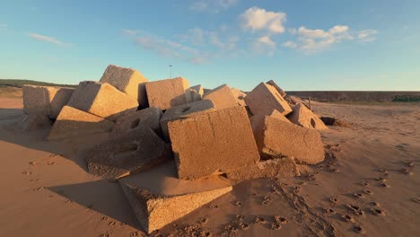 Stroll-among-concrete-blocks-and-cubes-on-the-beach,-coastal-defense-structures-half-buried-in-sand,-illuminated-by-low-angle-sunlight