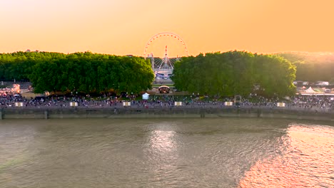 Ferris-wheel-with-people-walking-the-shoreline-of-Garonne-river-and-Monument-of-Girondins-during-wine-fair,-Aerial-approach-shot