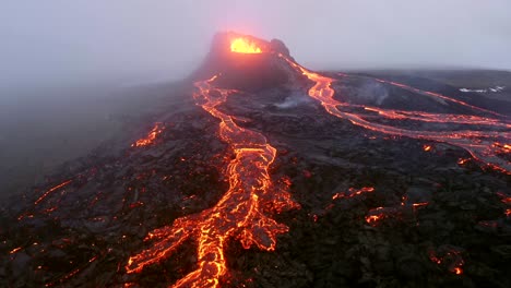 A-4K-drone-captures-aerial-cinematic-shots-of-an-active-volcano-ejecting-lava-rapidly,-followed-by-cascading-lava-amidst-misty-and-foggy-surroundings-in-Iceland