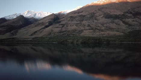 Fliegen-Sie-Bei-Sonnenuntergang-Zu-Den-Schneebedeckten-Bergen,-Wobei-Die-Spiegelungen-Im-See-Eine-Perfekte-Symmetrie-In-Queenstown,-Neuseeland-Erzeugen