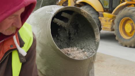 Male-construction-worker-filling-electric-cement-mixer-gravel-spinning-slow-motion-close-up