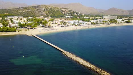 Retreating-drone-shot-of-the-beachfront-of-Playa-del-Port-de-Pollenca,-also-known-as-Puerto-Pollensa-beach-resort,-located-in-the-island-of-Mallorca,-Spain