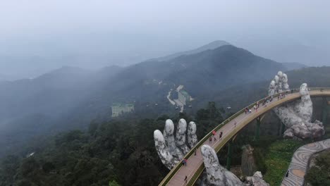Drone-aerial-view-in-Vietnam-vertical-descend-over-the-golden-bridge-held-by-giant-stone-hands-on-the-top-of-a-mountain-covered-by-green-trees-and-fog-with-people-walking-by-in-Ba-Na-Hills
