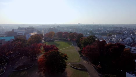 A-vibrant-city-park-with-autumn-foliage-at-sunset,-skyline-in-the-backdrop,-aerial-view