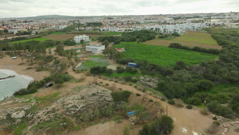 Vista-Aérea-De-Una-Zona-Costera-Que-Combina-Paisajes-Naturales-Y-Urbanos,-Con-Terreno-Rocoso-Que-Conduce-A-Campos-Agrícolas-Y-Una-Serie-De-Edificios-Con-Una-Ciudad-En-La-Distancia.