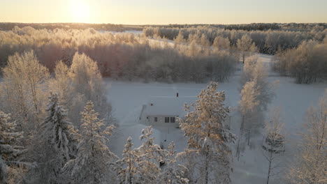 Remote-modern-Nordic-home-in-winter-woodland-with-glowing-sun-above,-aerial-view