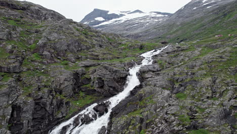 Aerial-shot,-tracking-up-from-the-top-of-the-large-Stigfossen-waterfall-to-reveal-a-large-snow-capped-mountains-and-a-wide-valley-in-the-distance