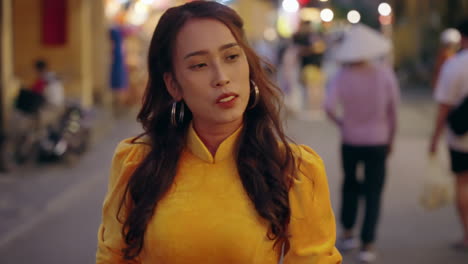 Young-woman-in-vibrant-yellow-Ao-Dai-strolls-through-Hoi-An's-bustling-evening-market,-close-up