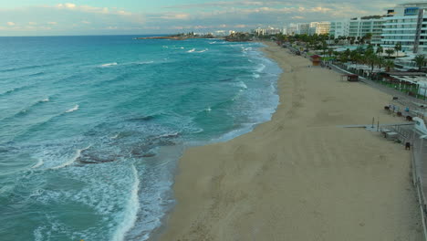 Aerial-view-of-the-vibrant-Protaras-coastline-in-Cyprus,-golden-sandy-beach-with-frothy-waves-lapping-at-the-shore,-flanked-by-a-promenade-with-palm-trees-and-modern-resort-buildings