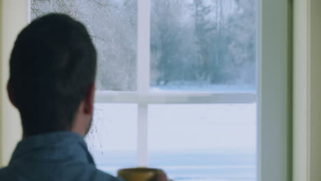 Young-man-drinking-morning-coffee-and-looks-through-window-to-snowy-landscape