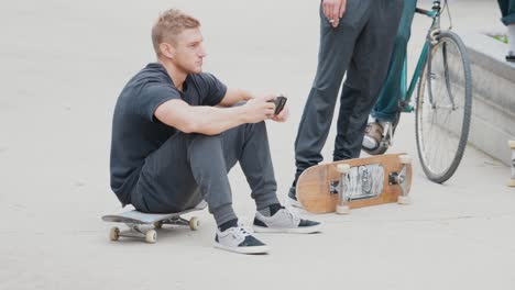 Ein-Junger-Mann-In-Schwarzer-Kleidung,-Der-Auf-Einem-Skateboard-Sitzt-Und-Sein-Telefon-Benutzt