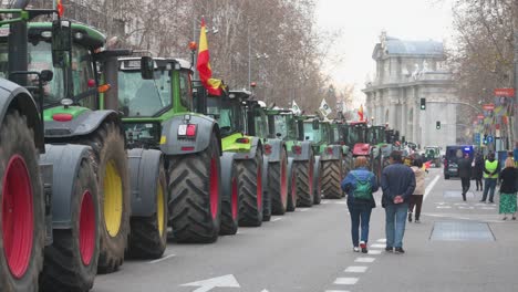 Pedestrians-walk-past-a-line-of-tractors-parked-on-the-street-during-a-farmer-strike-as-farmers-and-agricultural-unions-protest-against-unfair-competition,-agricultural-and-government-policies