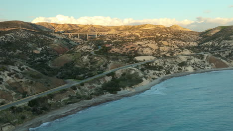 Scenic-view-near-Aphrodite's-Rock-with-a-winding-road-hugging-the-coastline,-flanked-by-hills-and-a-distant-bridge-against-the-backdrop-of-a-golden-sunset
